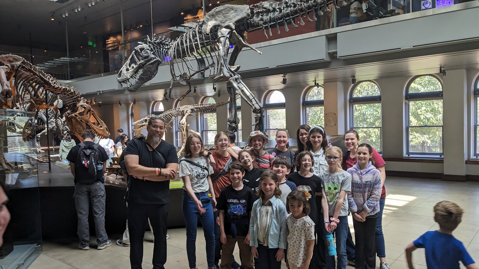 Group of kids and adults posing for a photo inside a museum beside large dinosaur skeletons on display.