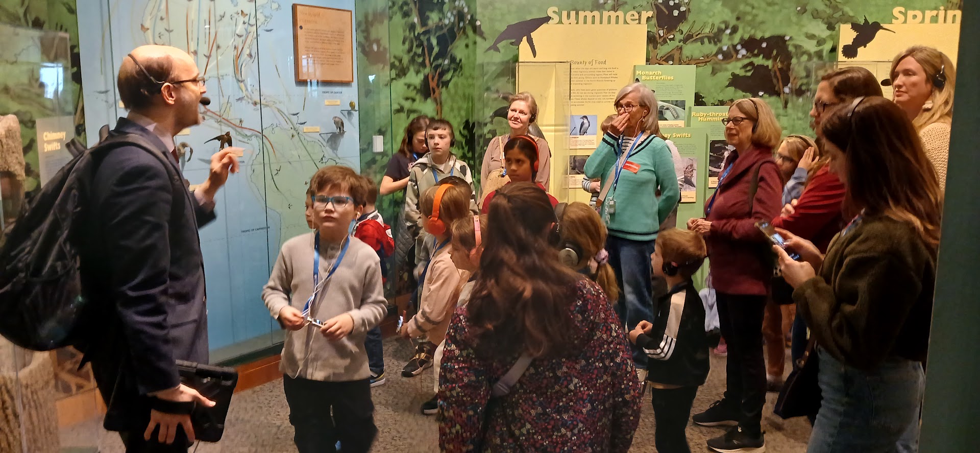 Museum guide giving tour to a group of children and adults in front of an exhibit wall with maps and text panels.